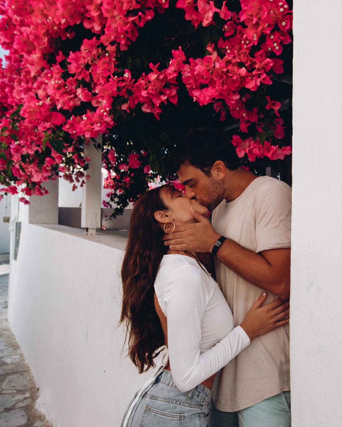 A couple kissing beneath bright pink flowers beside a white wall