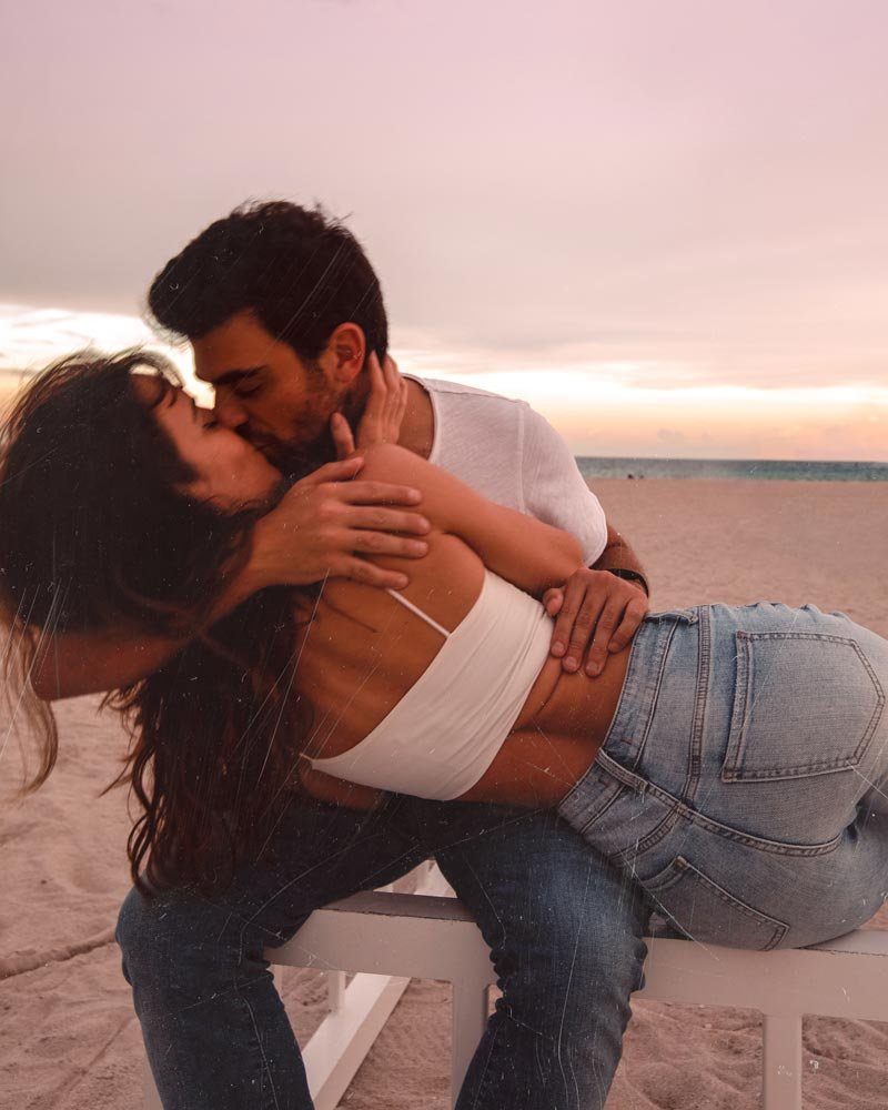 A couple sharing a kiss on a beach beneath a pink sunset sky